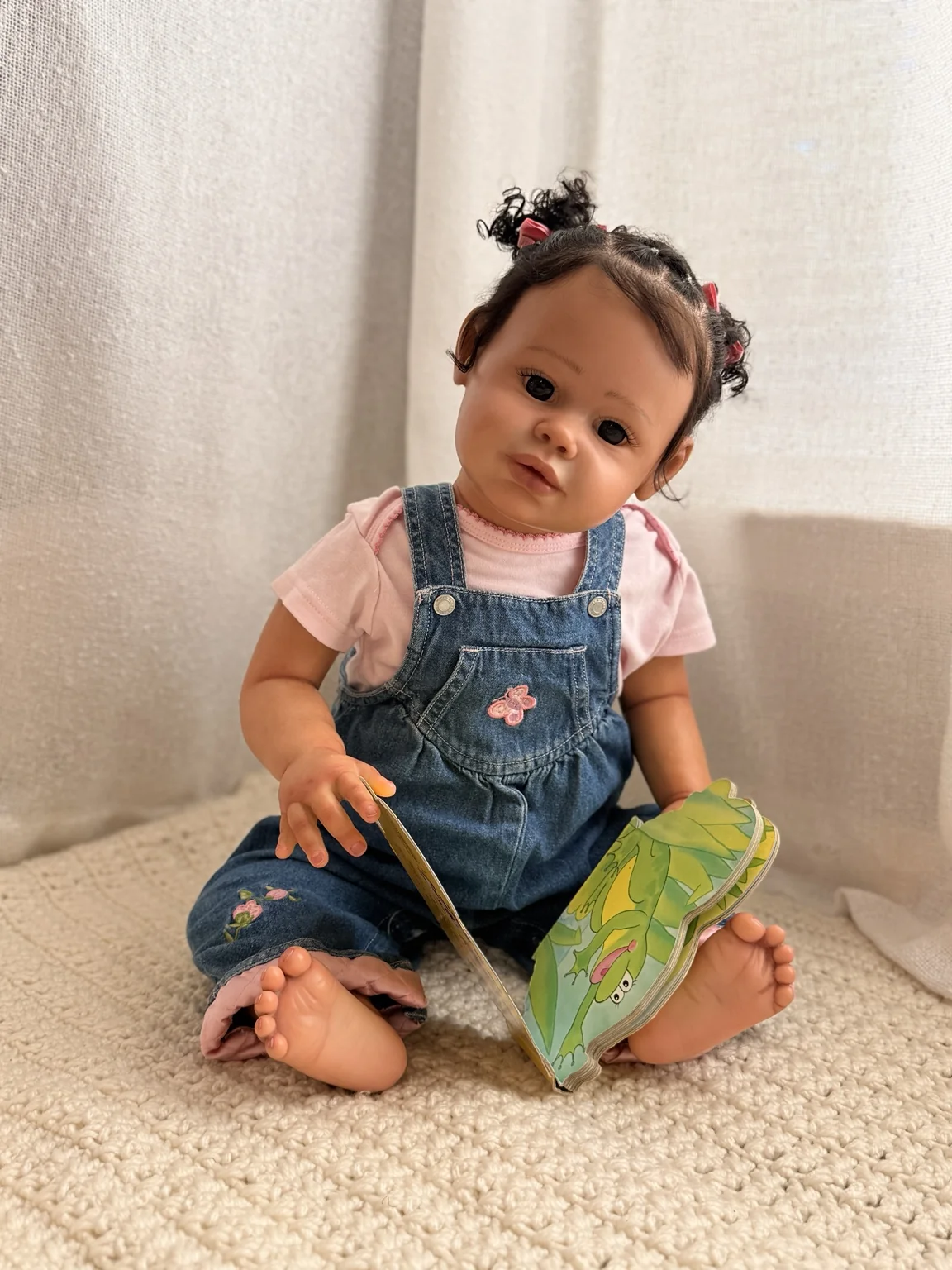Reborn doll in pink and embroidered denim overalls seated with an open picture book on a cream blanket